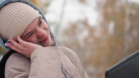 close view of light skin lady in beige jacket and ribbed beanie smiling while talking on mobile phone with blue case, supporting phone with shoulder, blurred outdoor background in autumn season