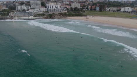 Surfing-Tourists-In-Blue-Ocean-With-Icebergs-Swimming-Pool-In-Background-At-Bondi-Beach,-Eastern-Suburbs,-Sydney,-NSW-Australia