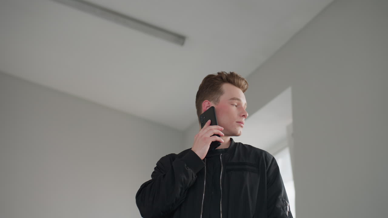 Young man in black jacket holds smartphone to ear speaking with boss, turns head to look outside bright window overlooking cityscape, neutral minimalist interior
