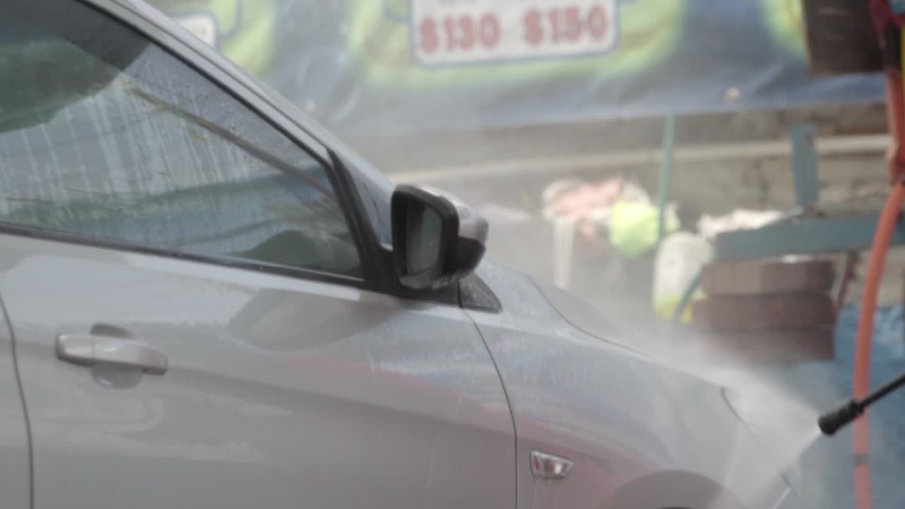A person pressure washing the front side of a silver car at an outdoor car wash station