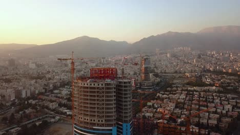 Girando-Alrededor-De-La-Torre-En-Construcción-Toma-Aérea-De-Drones-En-Teherán-Irán-Edificios-Del-Paisaje-Urbano-En-Segundo-Plano-En-El-Atardecer-Y-La-Montaña-En-El-Paisaje-Y-La-Contaminación-Del-Aire-Visible-En-El-Horizonte