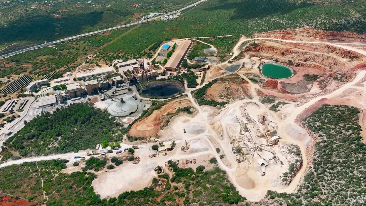 Aerial view shows a cement production facility and limestone quarry. Heavy machinery operates in rock extraction and processing, while conveyor belts transport materials throughout the site.