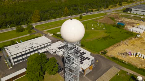 aerial drone revealing footage of a large white weather measuring radar dome or radome during a sunny summer day in a countryside. the radar ball is on a large tower above a weather station building