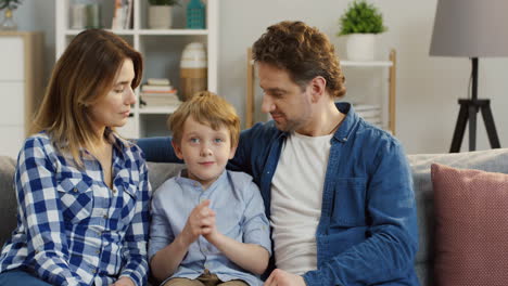 Portrait-Shot-Of-The-Young-Attractive-Parents-Sitting-With-Their-Little-Cute-Son-On-The-Sofa-In-The-Living-Room,-Hugging-And-Smiling-To-The-Camera