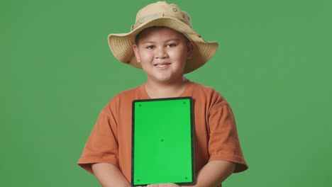 asian tourist boy with a hat and binoculars smiling and showing green screen tablet. boy researcher examines something on the green screen background, travel tourism adventure concept, close up