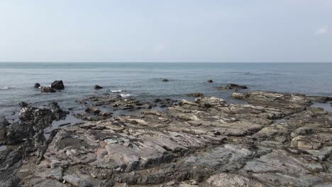 Aerial-flyover-of-rugged-shattered-bedrock-on-calm-sea-beach,-Thailand