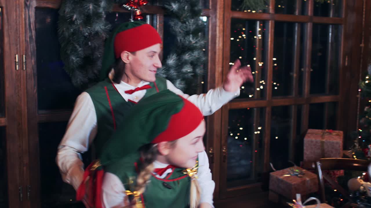 A girl and a boy Christmas elves are dancing near the window in a room with New Year decorations