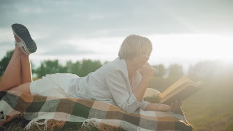 thoughtful woman reading book with hand on chin, lying on hay bale in vast open field, checkered blanket adds coziness while trees line the distant horizon under warm natural sunlight