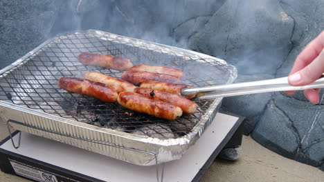 person turning sausages on a small instant bbq pit at the beach, between the rocks, in summertime