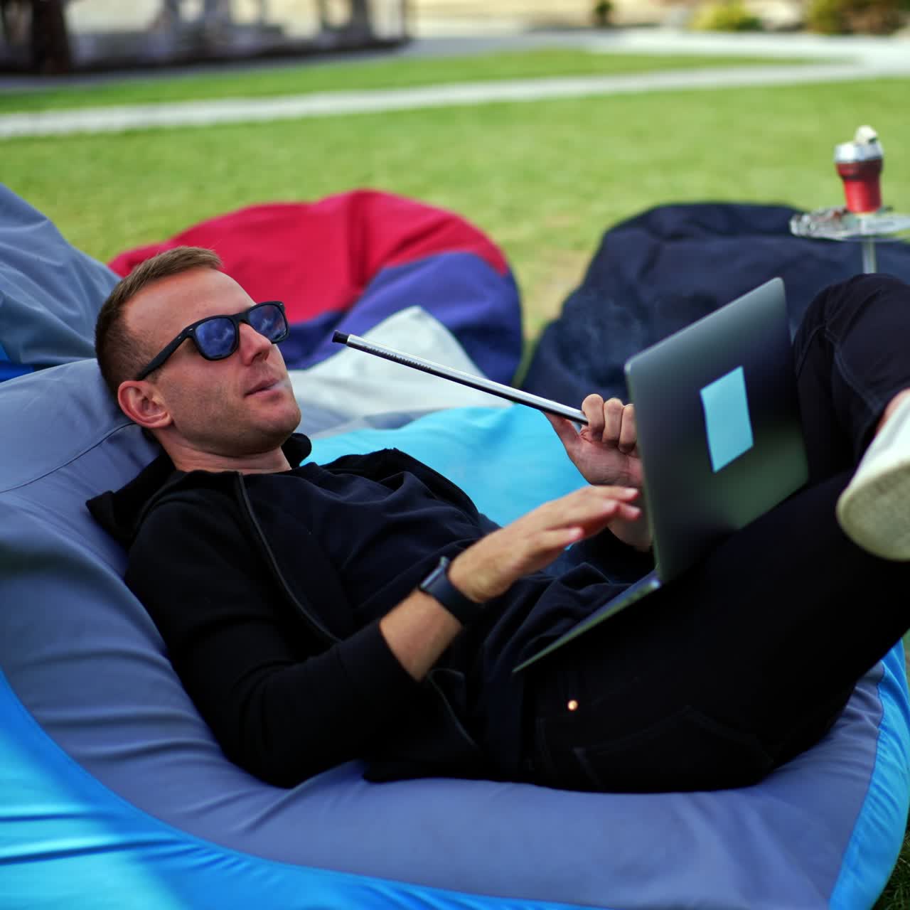 Mid-aged man in black clothes and sunglasses lies in bean bag chair outdoors. Resting male smoking hookah and looking at his laptop screen