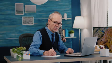 hombre jubilado multitarea leyendo en una computadora portátil y escribiendo en un cuaderno