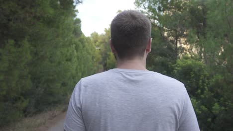 the back of a man dressed in basic gray t-shirt, who walks along a rural road surrounded by nature and trees on the sides of the image