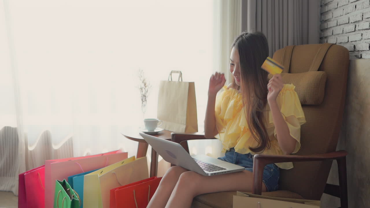 예쁜 아시아 소녀가 집에서 소파에 앉아 완료 후 즐기는 최신 노트북을 사용하여 은행 카드를 들고 온라인 결제를 하고 있습니다.