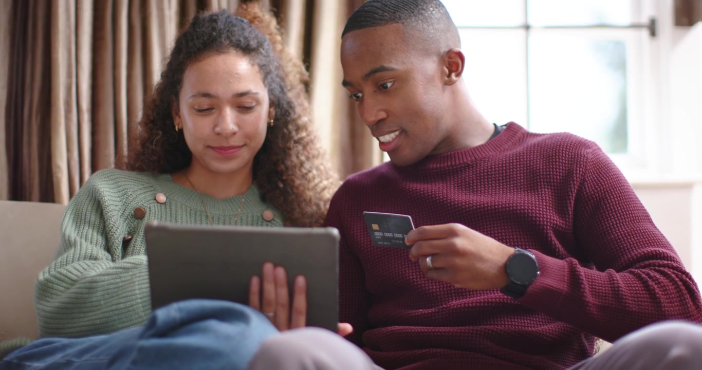 Diverse couple sitting couch, focus shifting from blue mug, checking tablet and entering card info