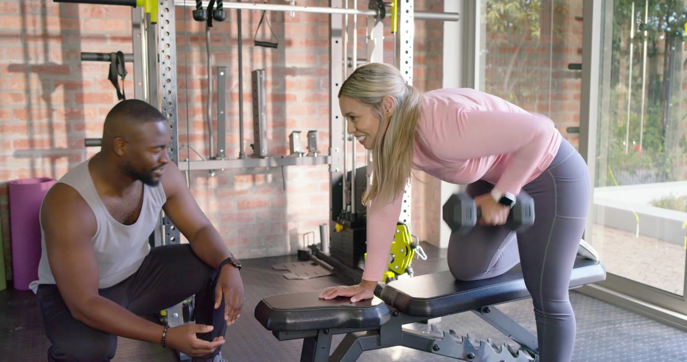 African American coach cueing as female client doing single-arm dumbbell rows on bench in home gym
