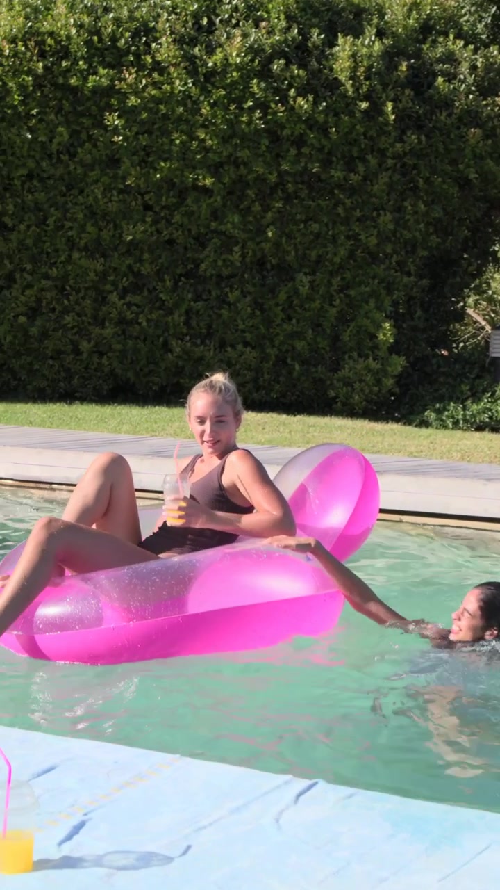 Vertical video: Splashing woman in swimwear swimming to pink float to join pal at pool, copy space