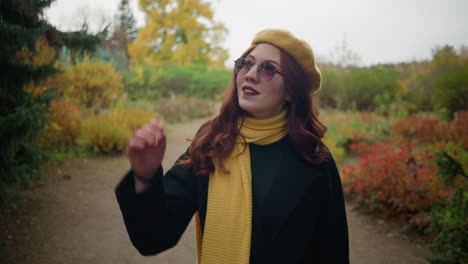 white lady in a yellow beret and matching scarf adjusts her red wavy hair while taking an autumn stroll, looking around with a blurred building in the distant background