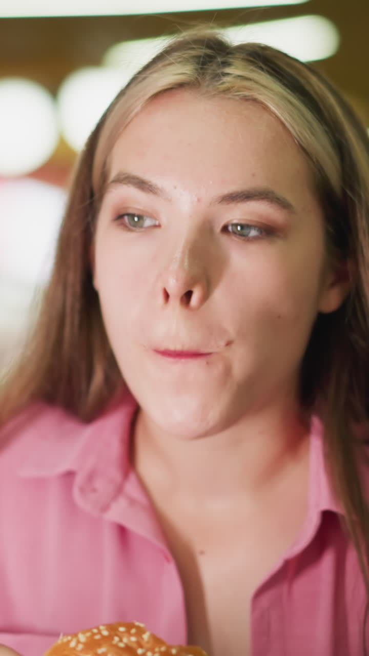 dama de vestido rosa toma un bocado de hamburguesa, masticando lentamente mientras contempla el sabor, parece reflexiva, saboreando la comida en un estado de ánimo relajado, el fondo tiene luces bokeh suaves