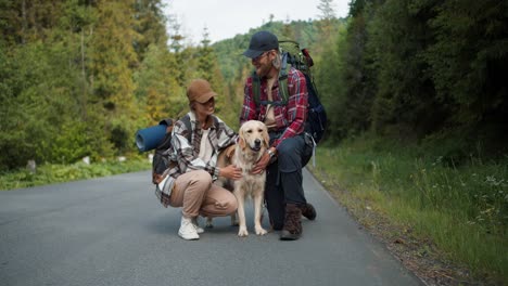 a guy and a girl tourists in special clothes for hiking stand near the road and stroke their light-colored dog. a couple of blondes against the backdrop of a green mountain forest