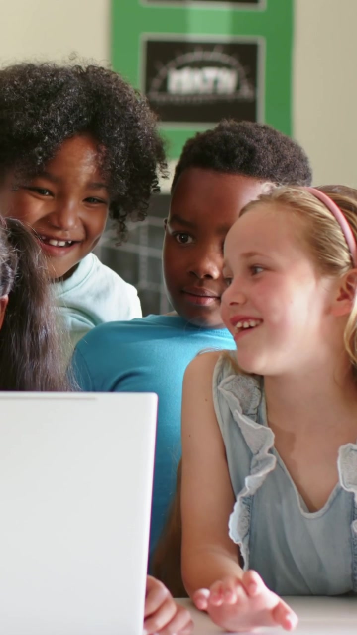 Vertical video: Tapping laptop, blonde girl guiding two classmates at school desk, watching screen