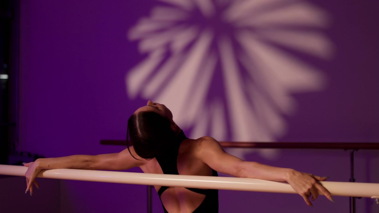 Graceful woman stretching her back and arms at the ballet barre in a dance studio with purple light