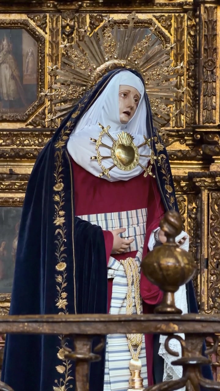 Náboženská socha v bohatě zdobeném rouchu stojí před zlaceným oltářem uvnitř katedrály.