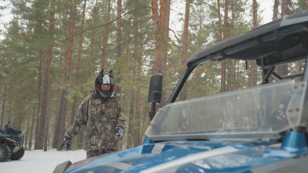 Persona in mimetica che si avvicina a un quad blu in una pineta innevata, casco e occhiali visibili. Ispeziona parabrezza e specchietti, valutando il veicolo prima di una corsa estrema, sotto una luce atmosferica tesa e il freddo.