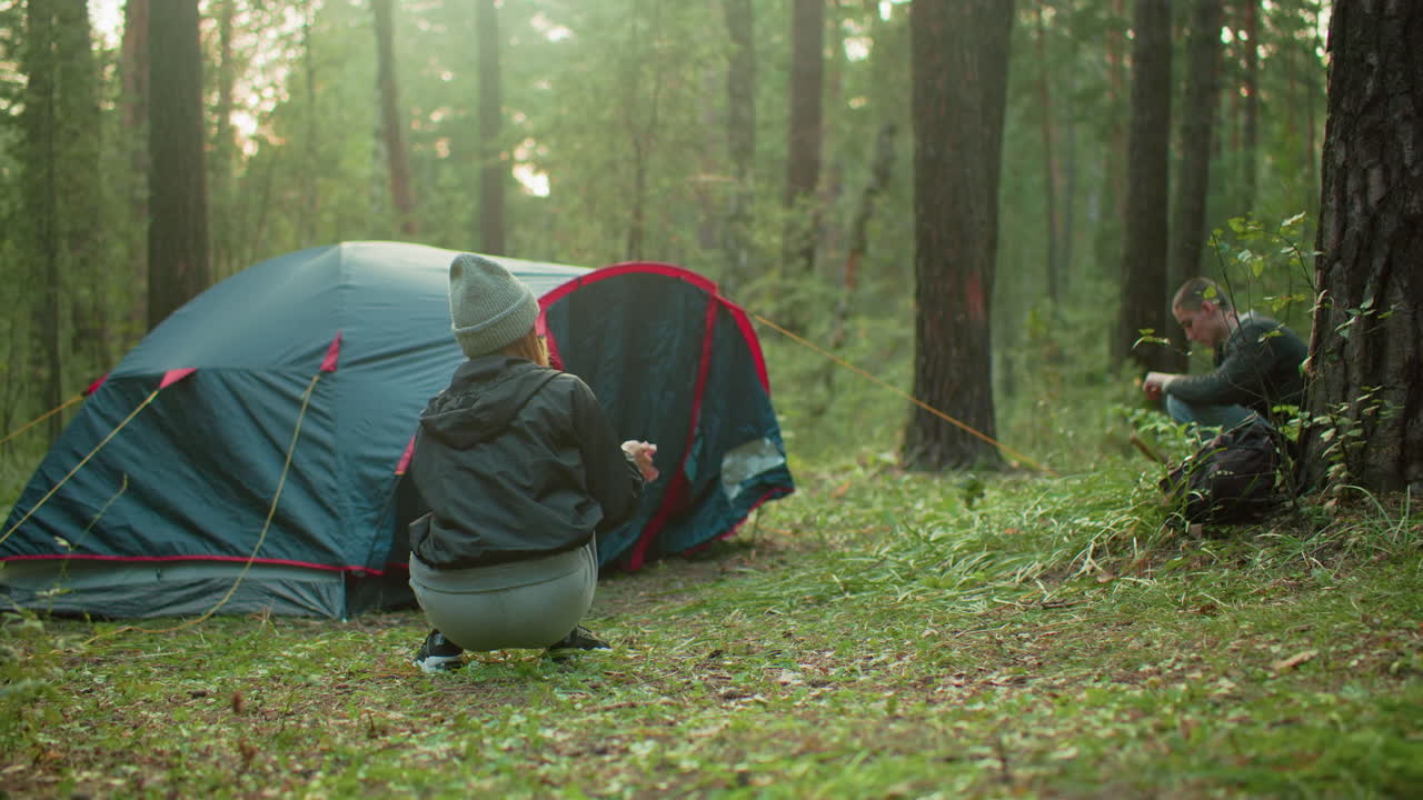 Woman dusts hands after completing tent setup while man rises from pegging final rope, both surrounded by forest trees in soft sunlight, completing peaceful morning