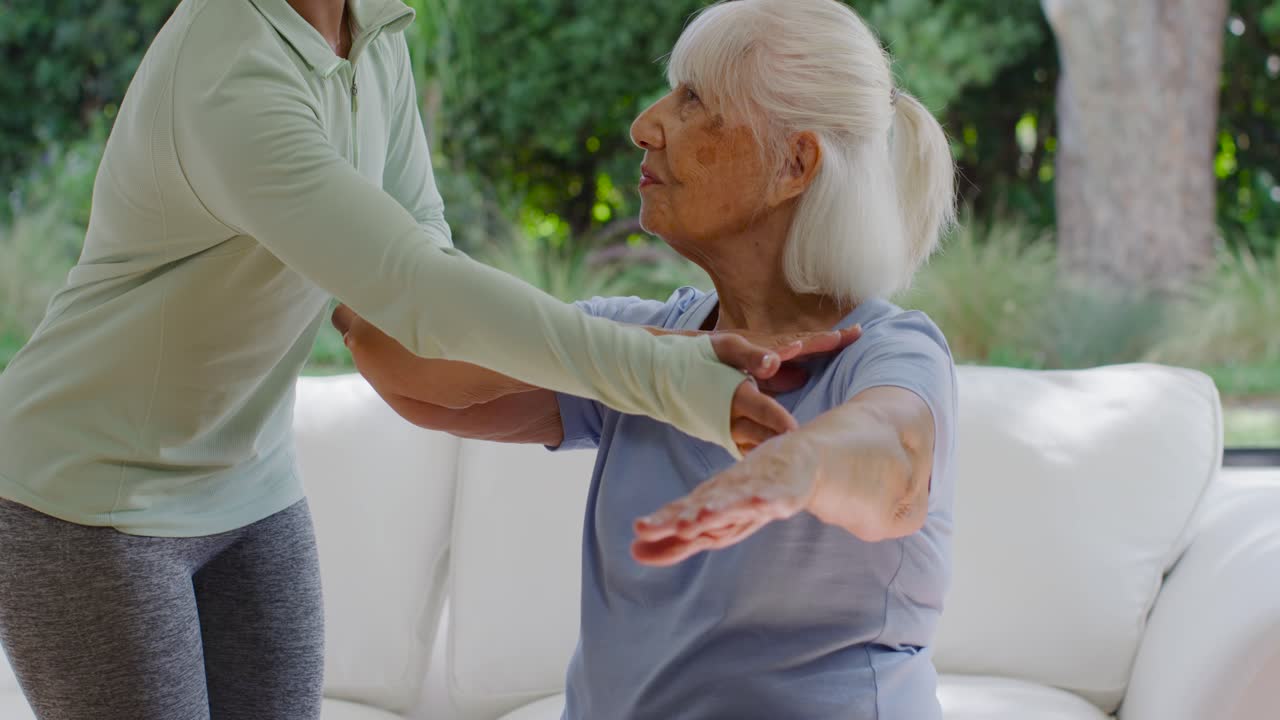 Adult African American caregiver showing arms, guiding senior raising arms for rehab on white sofa