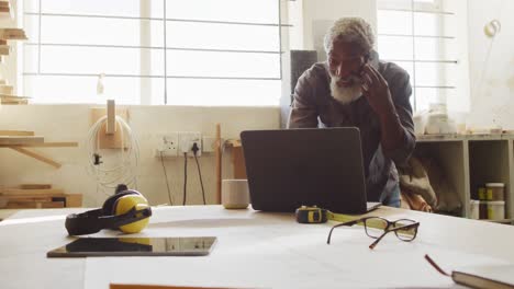 african american male carpenter talking on smartphone and using laptop in a carpentry shop