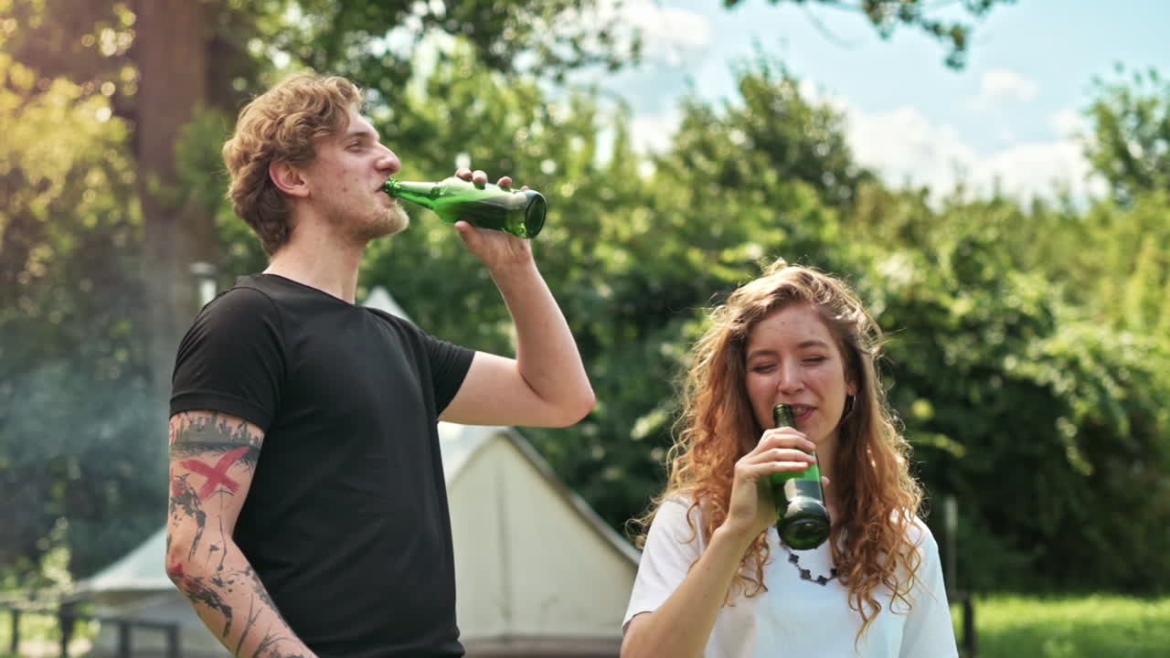 Young couple frying meat on the grill and drinking beer, laughing. Greenery around. Glamping. Slow motion