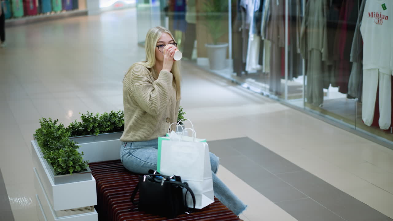 mujer profesional bebiendo café pensativa mientras está sentada cerca de plantas decorativas, el fondo presenta una tienda de ropa con maniquíes y compradores que pasan, creando una atmósfera de compras moderna