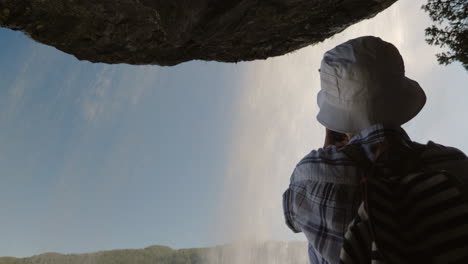 a woman standing under a waterfall steinsdalsfossen takes a photo above it hangs a rock the majestic