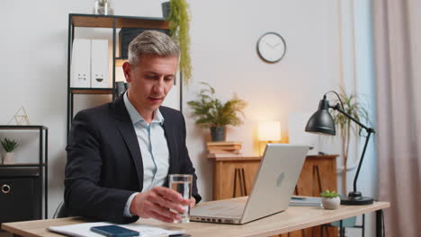 handsome young freelancer using laptop and drinking glass of water sitting at home office desk