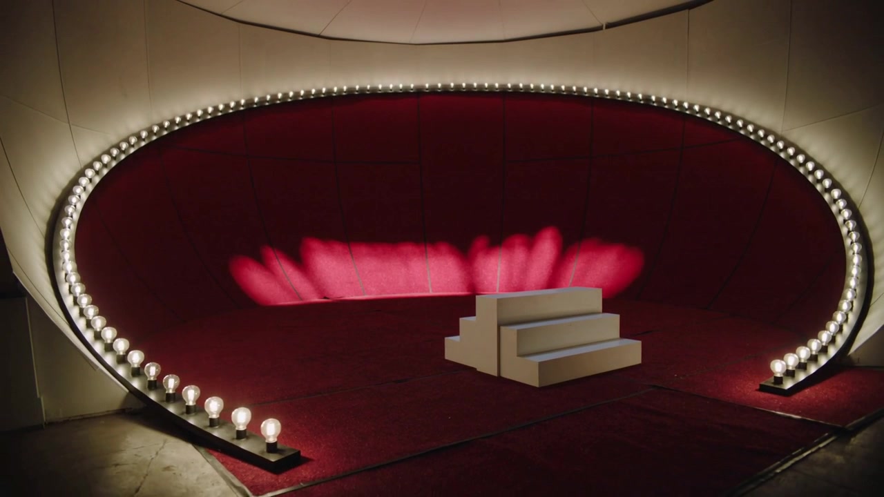 Projecting lights starting center forming red petal pattern on curved backdrop, with white platform