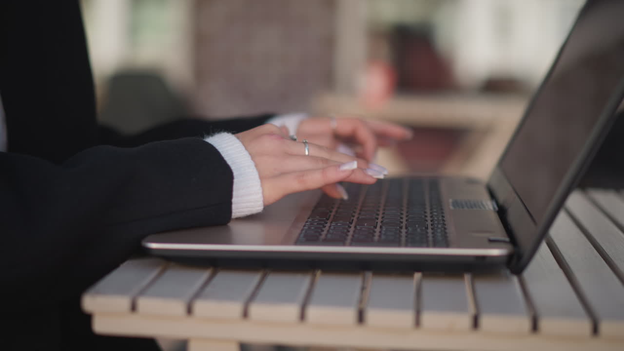 은 손을 가진 손이 노트북 키보드에서 타이핑하는 클로즈업, 은 반지와 아한 스웨터 팔꿈치로 장식되어 있으며, 흐릿한 배경에는 움직이는 인물이 있습니다.
