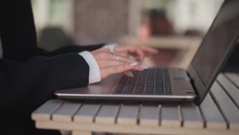 primer plano de manos con uñas pulidas escribiendo en el teclado de la computadora portátil, adornado con anillos de plata y cómodas mangas de suéter, con fondo borroso con figuras en movimiento