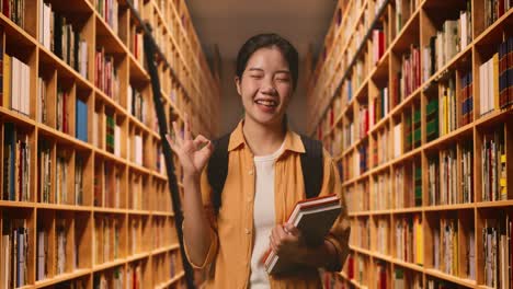 asian woman student with a backpack and some books smiling and showing okay gesture while standing in the library