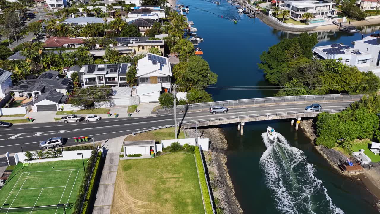 Drone footage captures a speedboat cruising through a canal beneath a road bridge in a sunny, upscale Gold Coast residential area with tennis courts