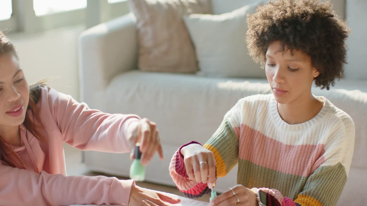 Diverse female friends selecting mint green polish and painting nails on small table in living room