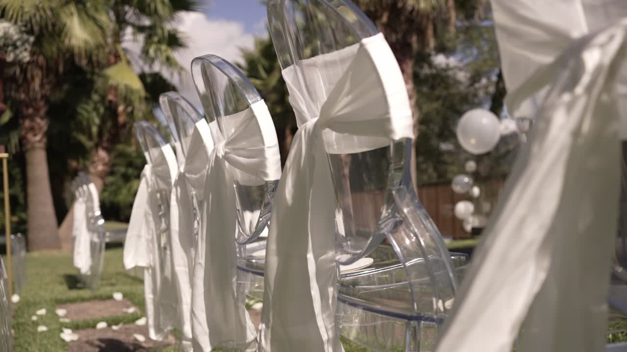 Clear chairs adorned with white cloth bows, set outdoors with palm trees in the background