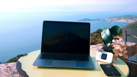 Close-up-tilt-up-of-business-laptop,smartphone-and-flower-on-table,-outdoors-with-beautiful-ocean-coastline-view-in-background