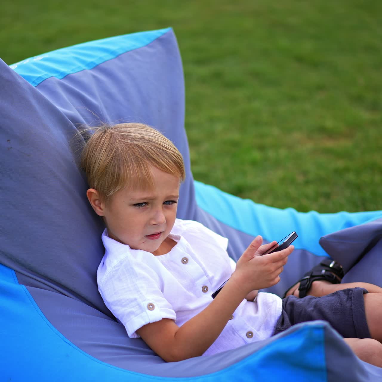 Blue-eyed blond boy of six years having rest outdoors. Lovely kid using smartphone sitting in bean bag chair on the green grass