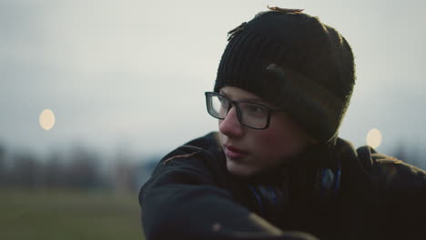 close-up of a young boy wearing glasses and a black beanie with grass particles on his head and headset on his neck, engaged in a discussion , with a blur background view