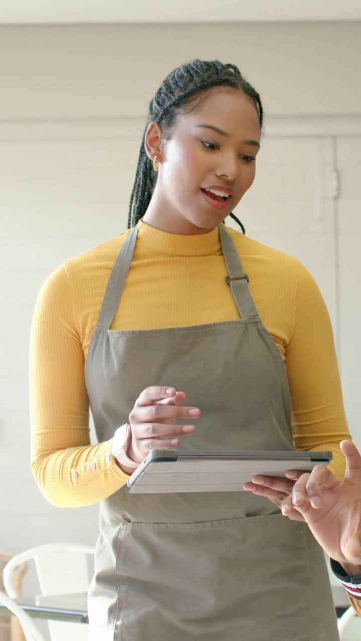 Vertical video: Tapping woman in gray apron using tablet at cafe after guest pointing, confirming