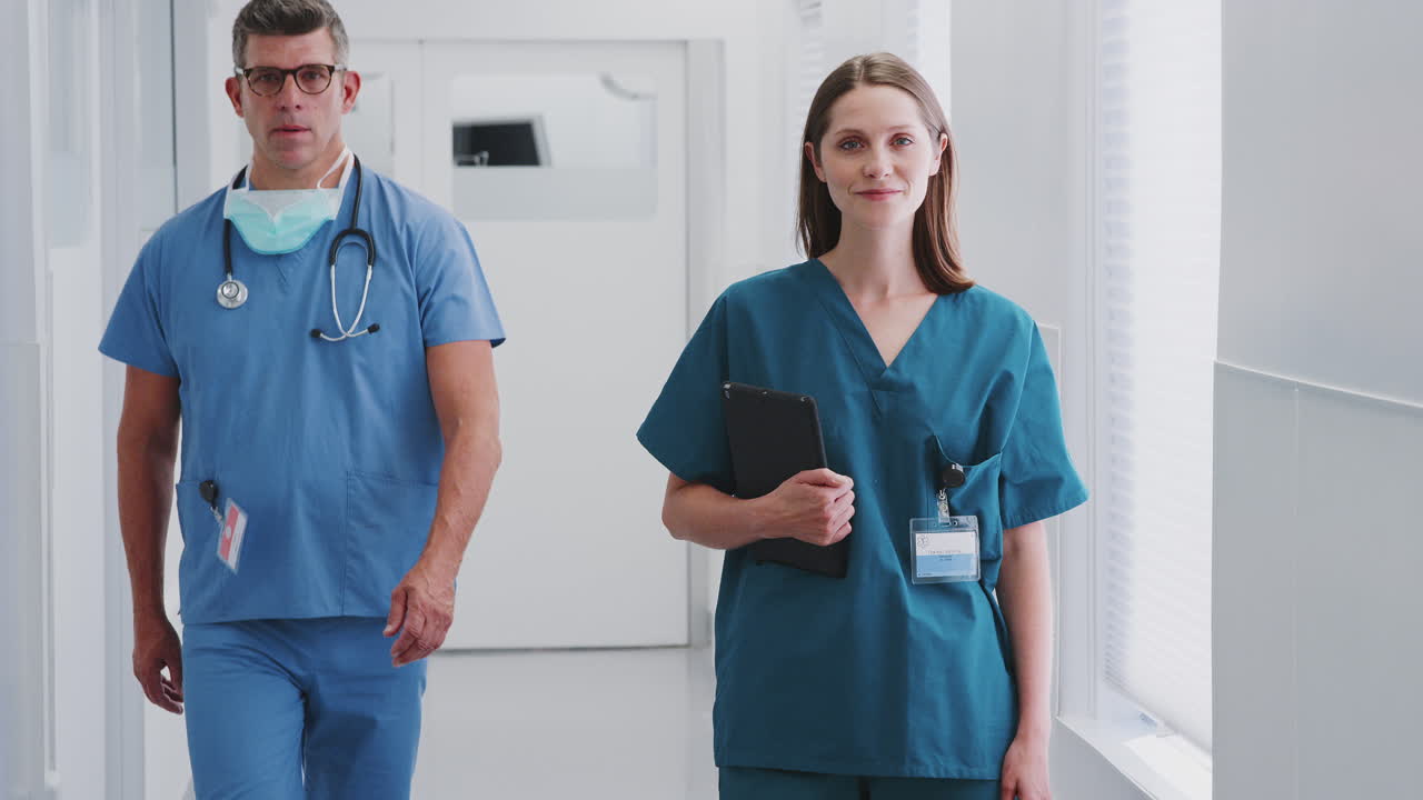 Portrait Of Smiling Female Doctor Wearing Scrubs In Busy Hospital Corridor Holding Digital Tablet