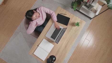 vista superior zoom out toma de una mujer estirándose mientras está sentada en el escritorio de madera escribiendo en una computadora portátil en casa