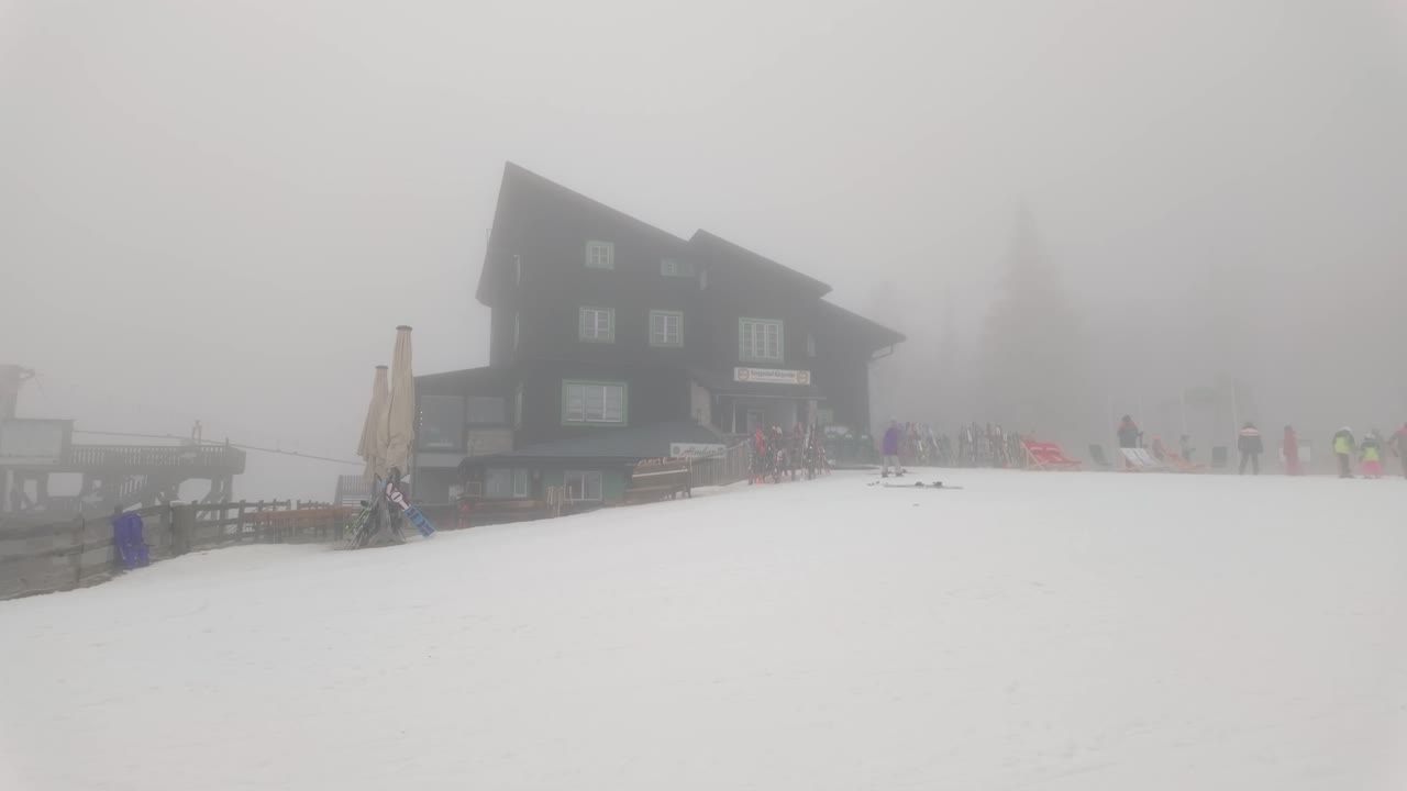 A cloud covered guest house located in a snowy mountain area, with people skiing nearby. The atmosphere is cold, and the scene is perfect for winter tourism in a ski resort.