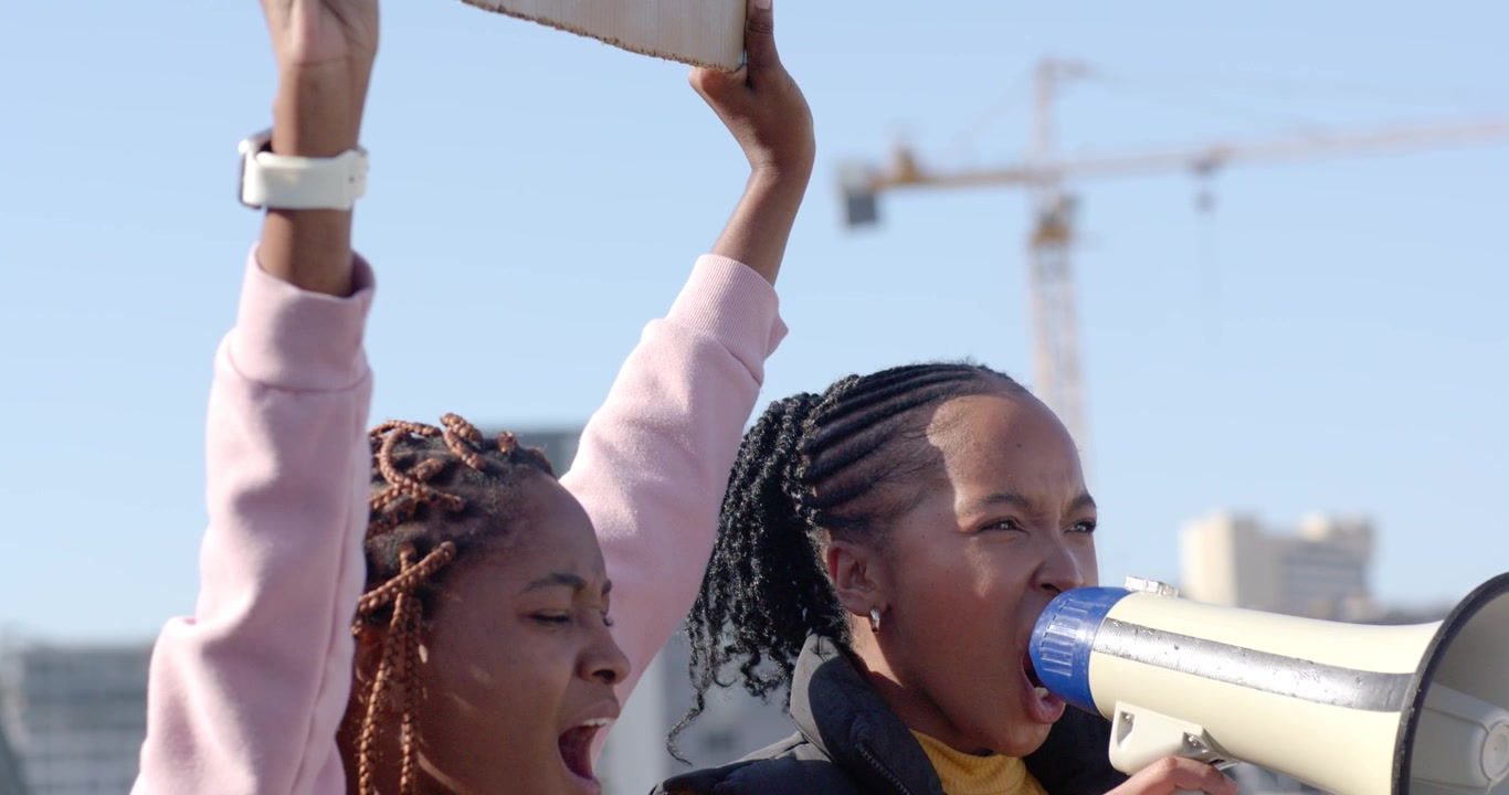 Two adult African women holding WOMEN sign raising arms shouting into megaphone in plaza protesting