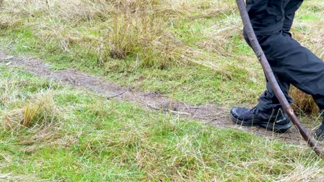 Person in outdoor gear walks with stick along grassy trail, natural daylight, steady camera movement